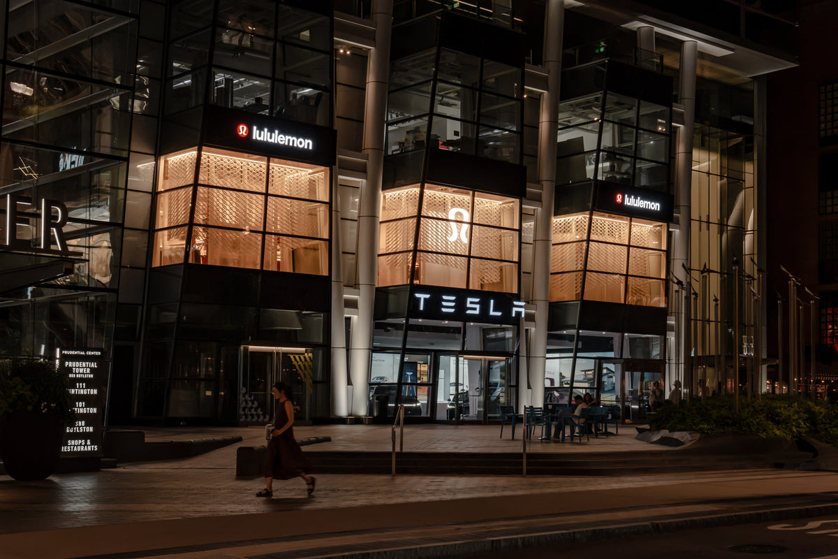 Wide night streetscape of lululemon’s three illuminated Prudential Center window bays with a pedestrian moving through the foreground in Boston.