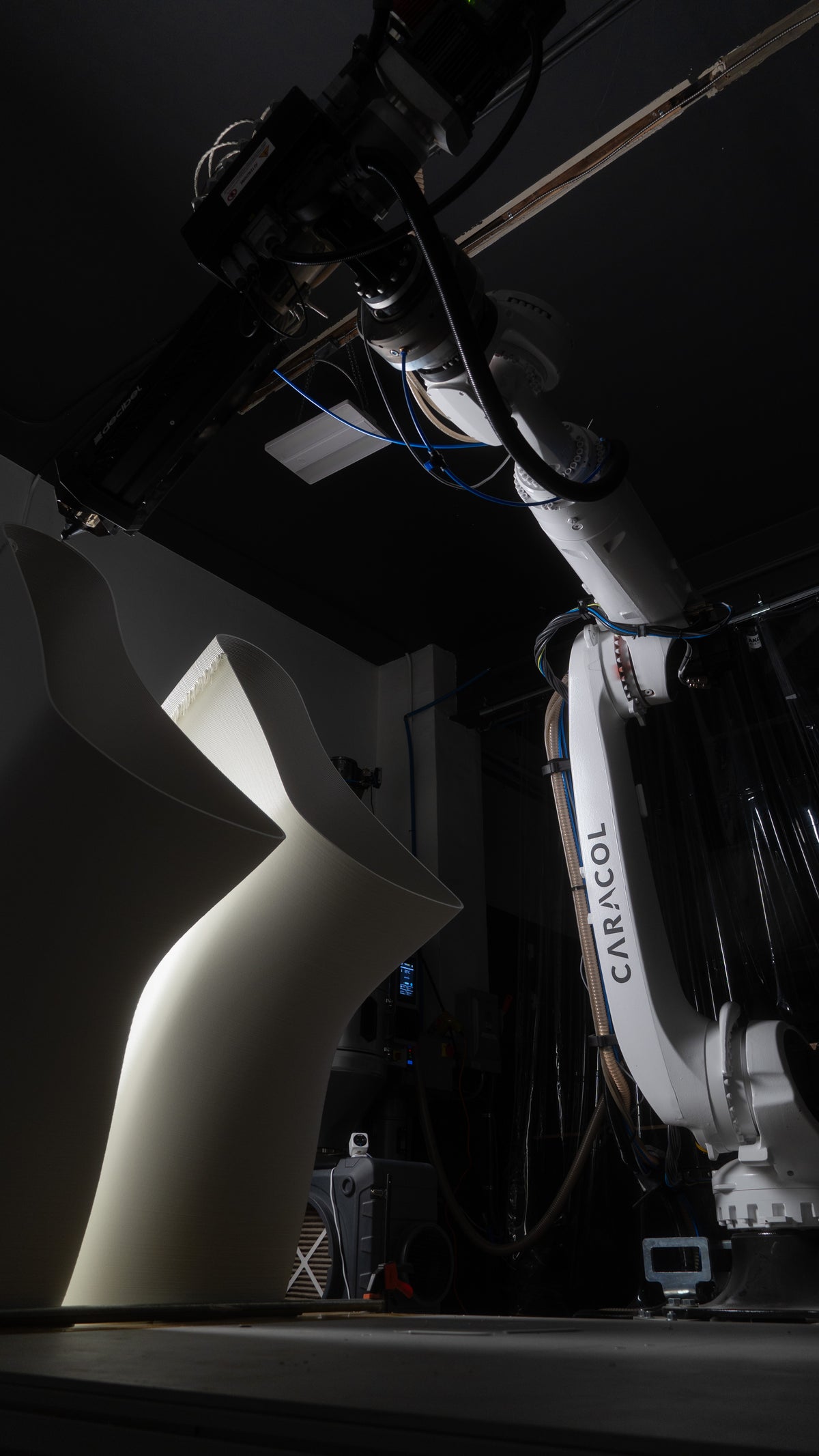 Robotic extrusion system beside a large curved white printed form in the Decibel workshop.